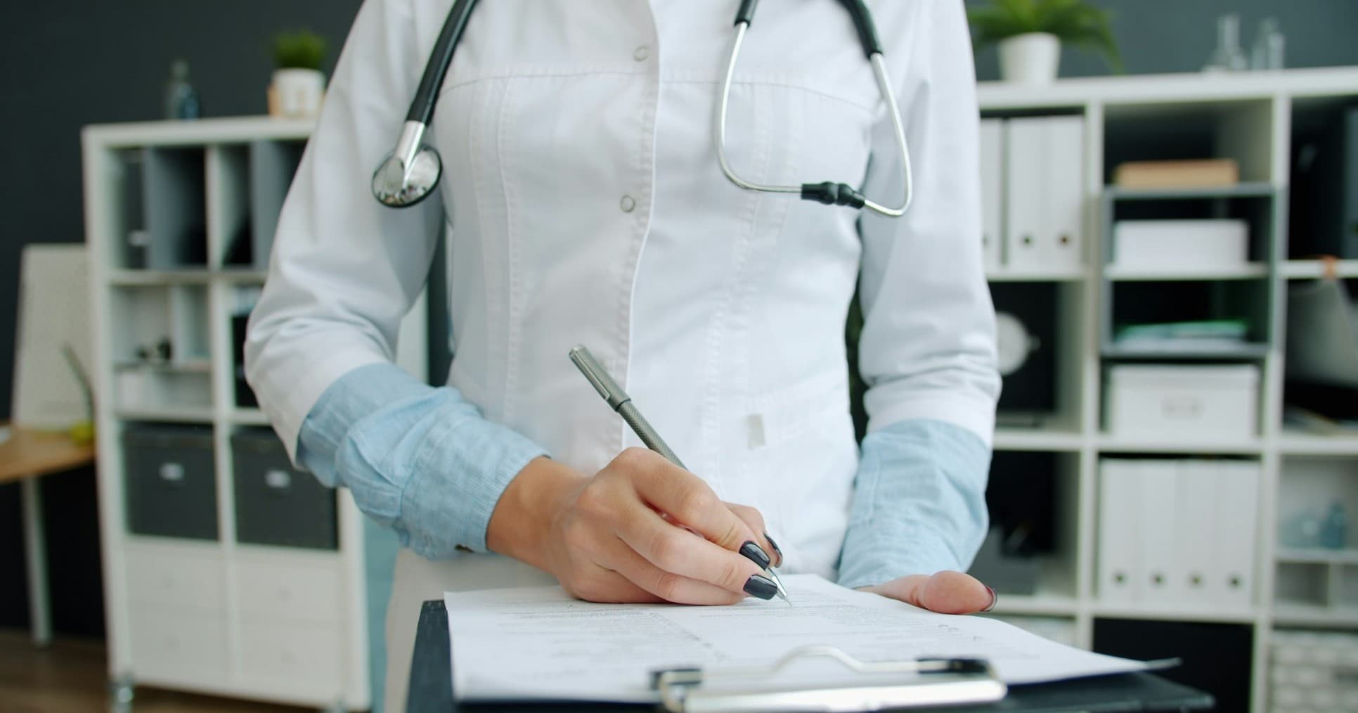 A clinician writes on a paper chart clipped to a clipboard in a medical office.