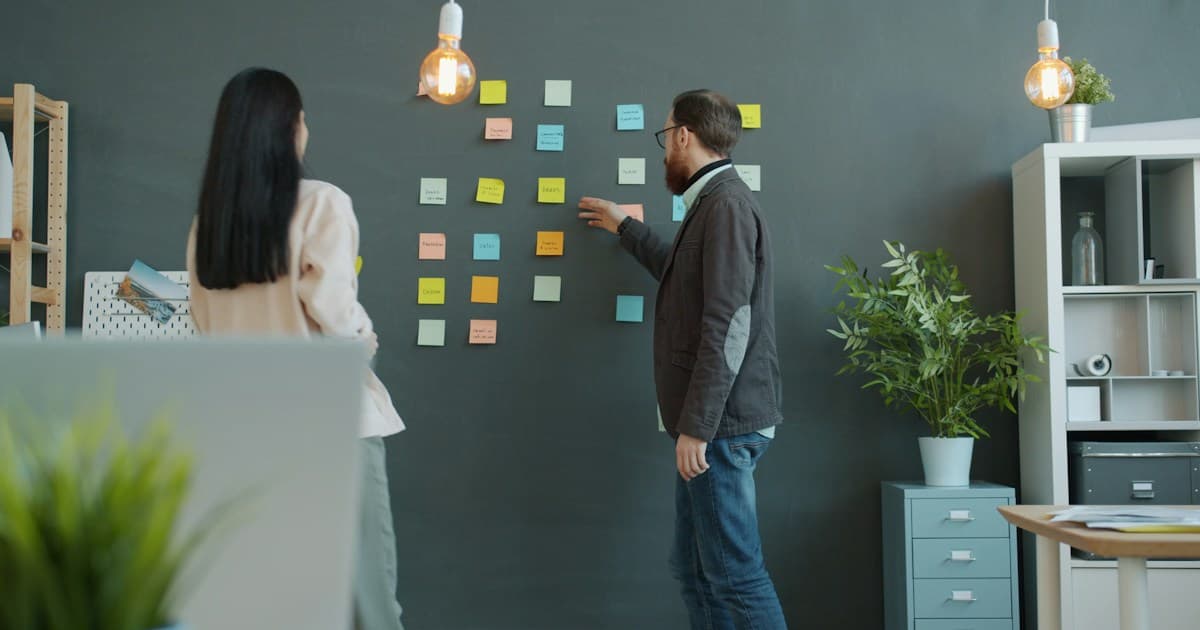 Two coworkers arrange sticky notes on a wall during a planning discussion.