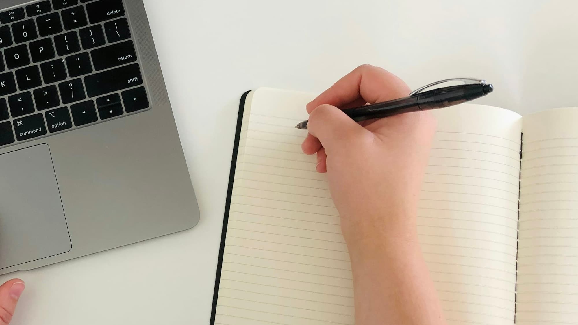 A person writes in an open notebook beside a laptop.