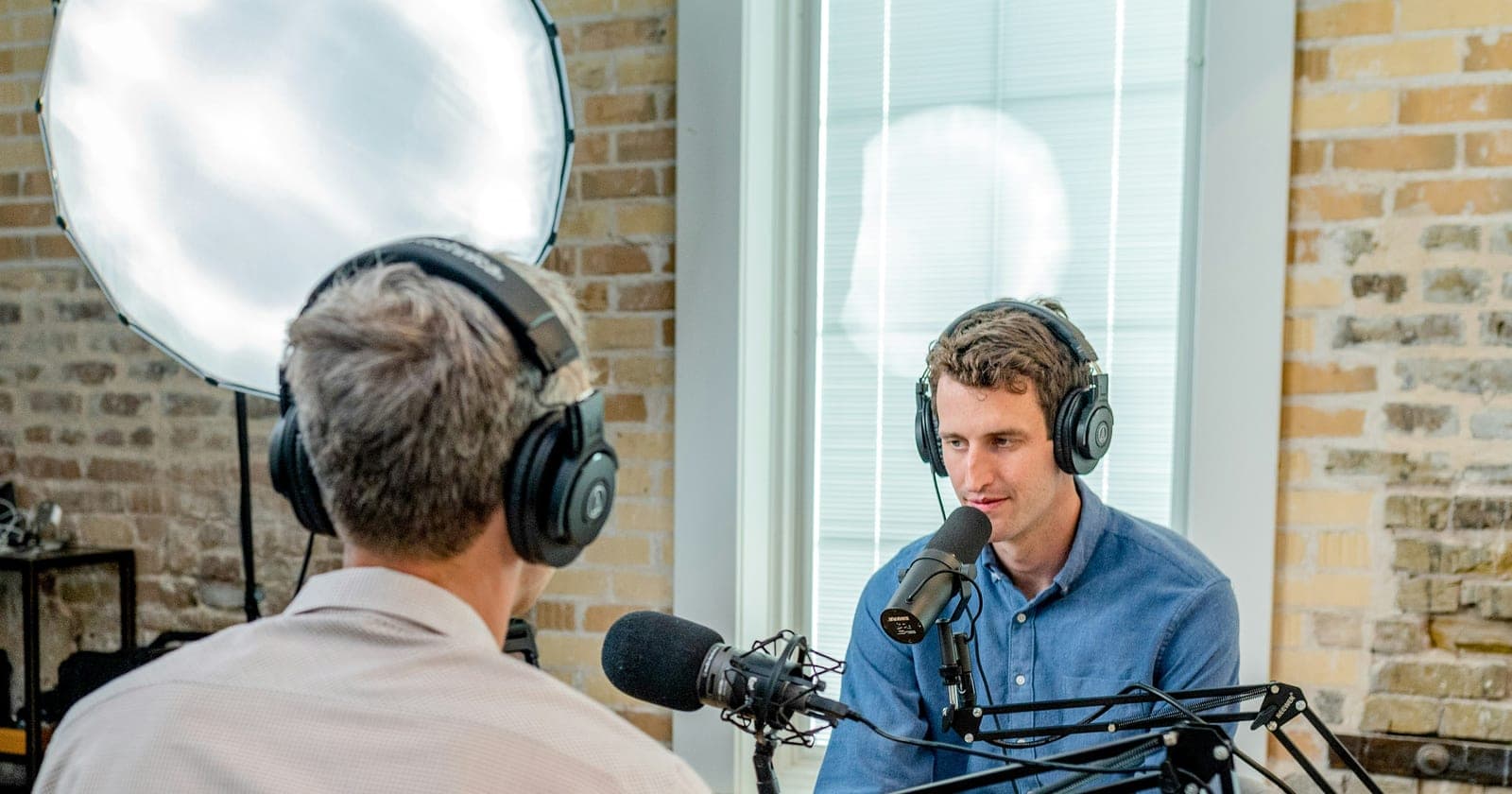 Two men wearing headphones speak across microphones in a podcast studio.