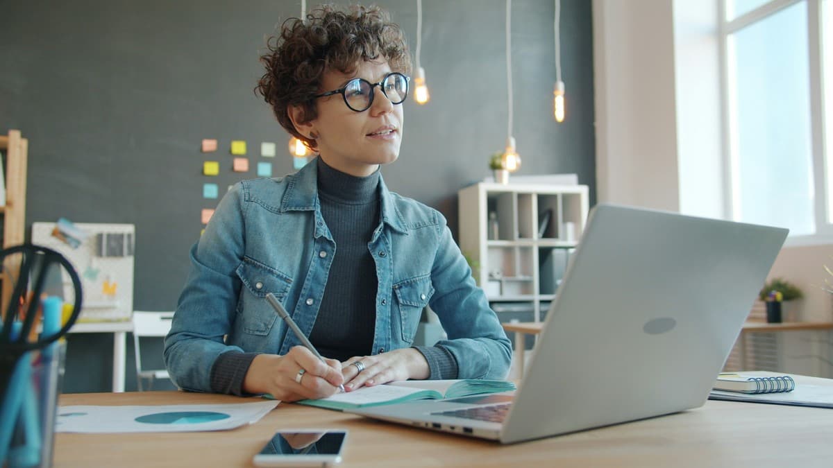 A woman writes in an open notebook at a desk beside a laptop.