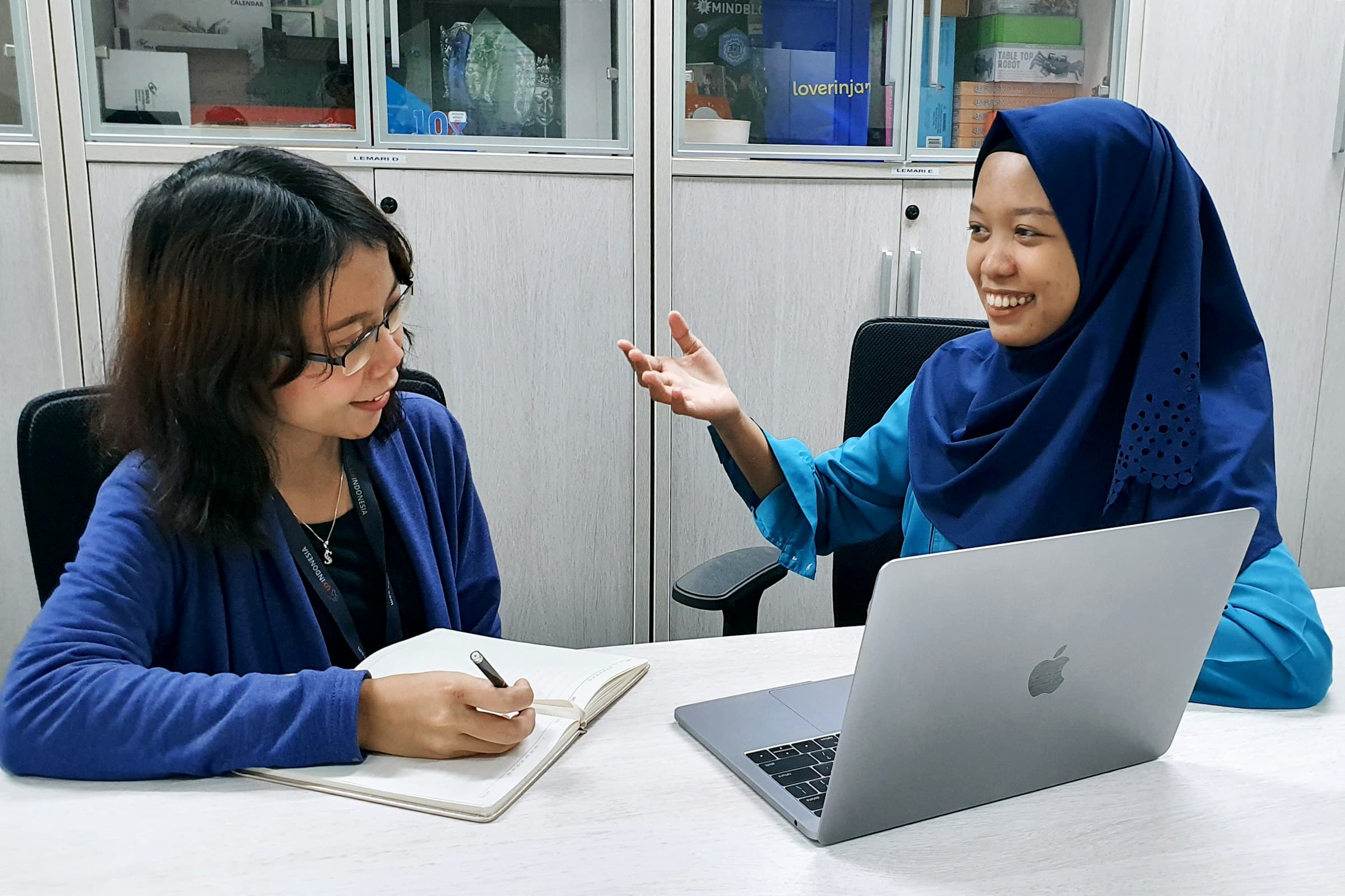 A researcher takes notes while listening to a participant beside an open laptop.