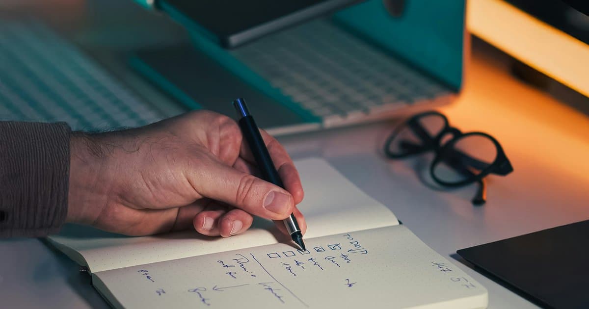 A hand marks a checklist in an open notebook beside a laptop and glasses.