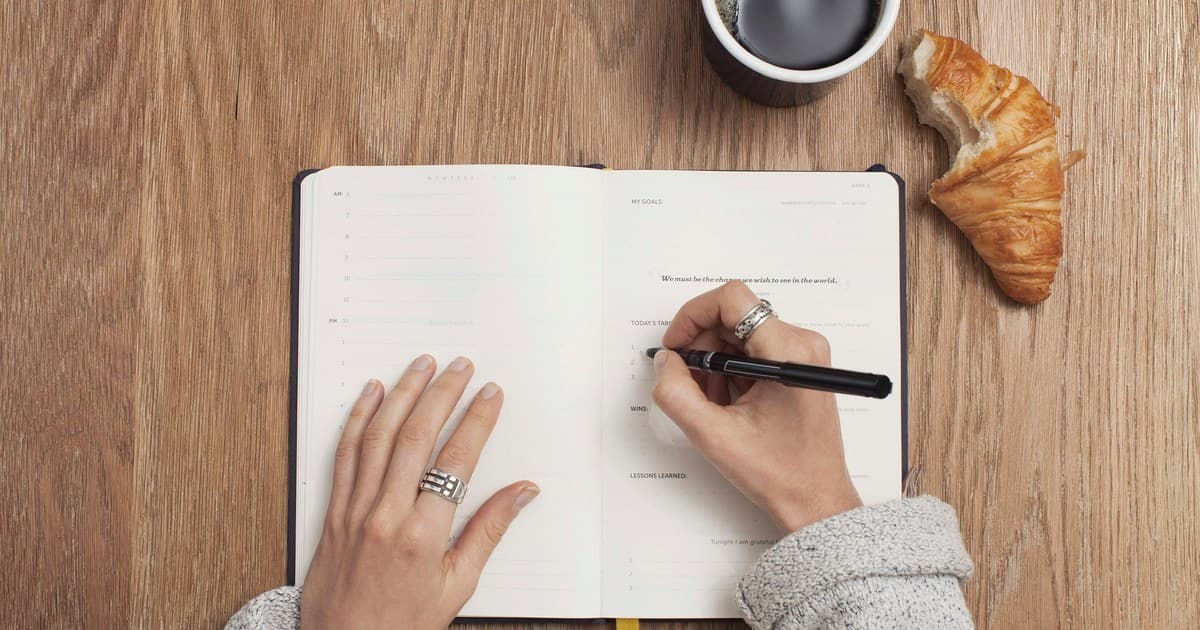 A person writes goals and reflections in an open journal at a wooden table.