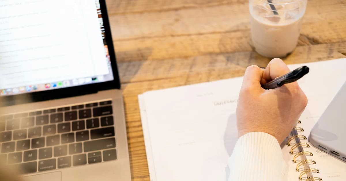 A person writes in a notebook beside an open laptop.