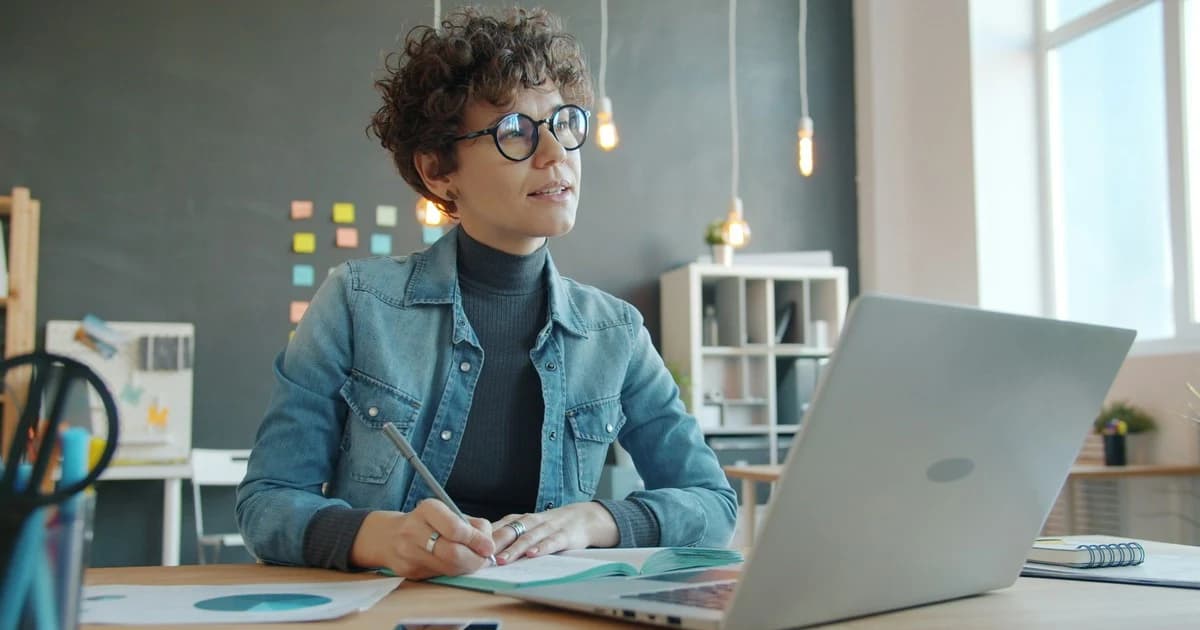 A woman writes in an open notebook beside a laptop.
