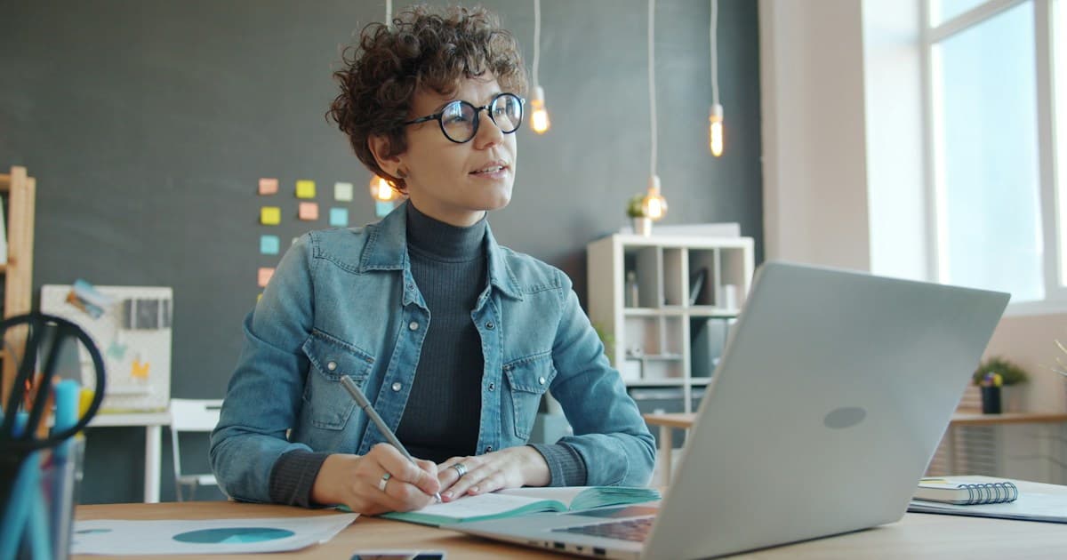 A researcher writes in a notebook beside a laptop at a desk.