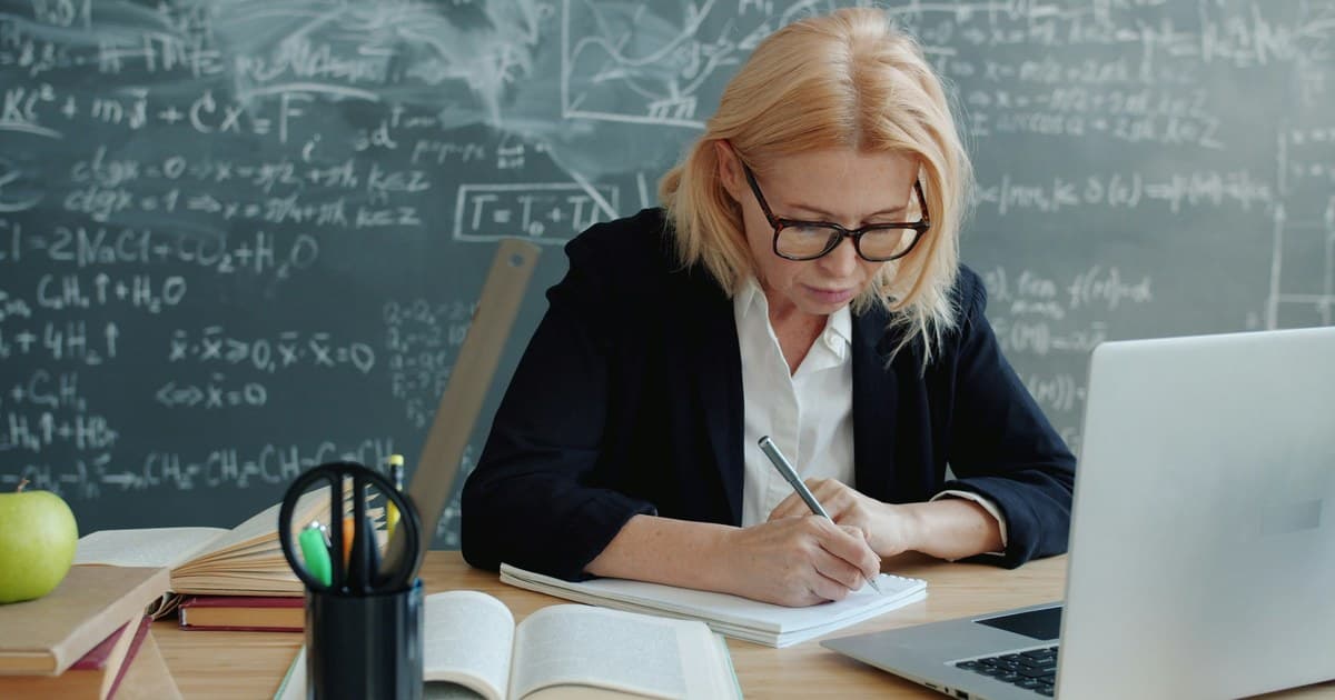 A woman writes in a notebook beside books and a laptop.
