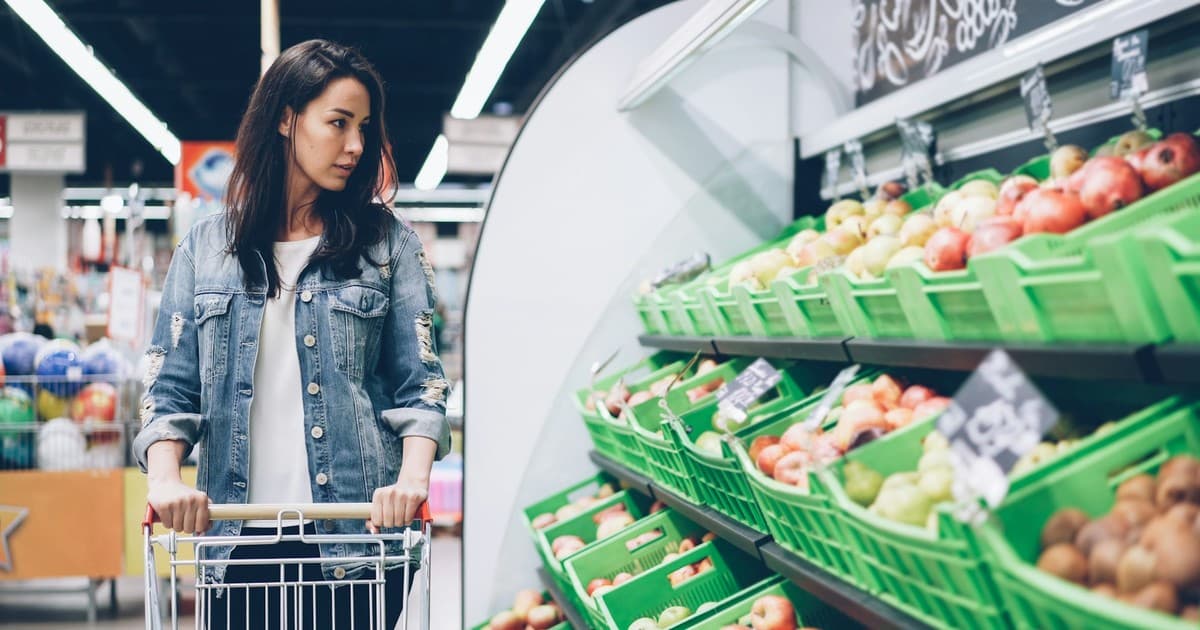 A woman selects produce from a supermarket display beside a shopping cart.