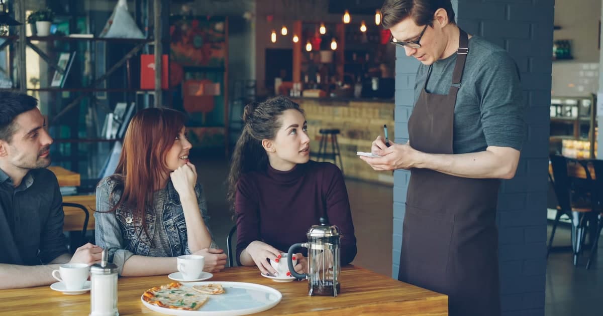 A waiter writes in a small order pad while three cafe customers look up and talk with him.