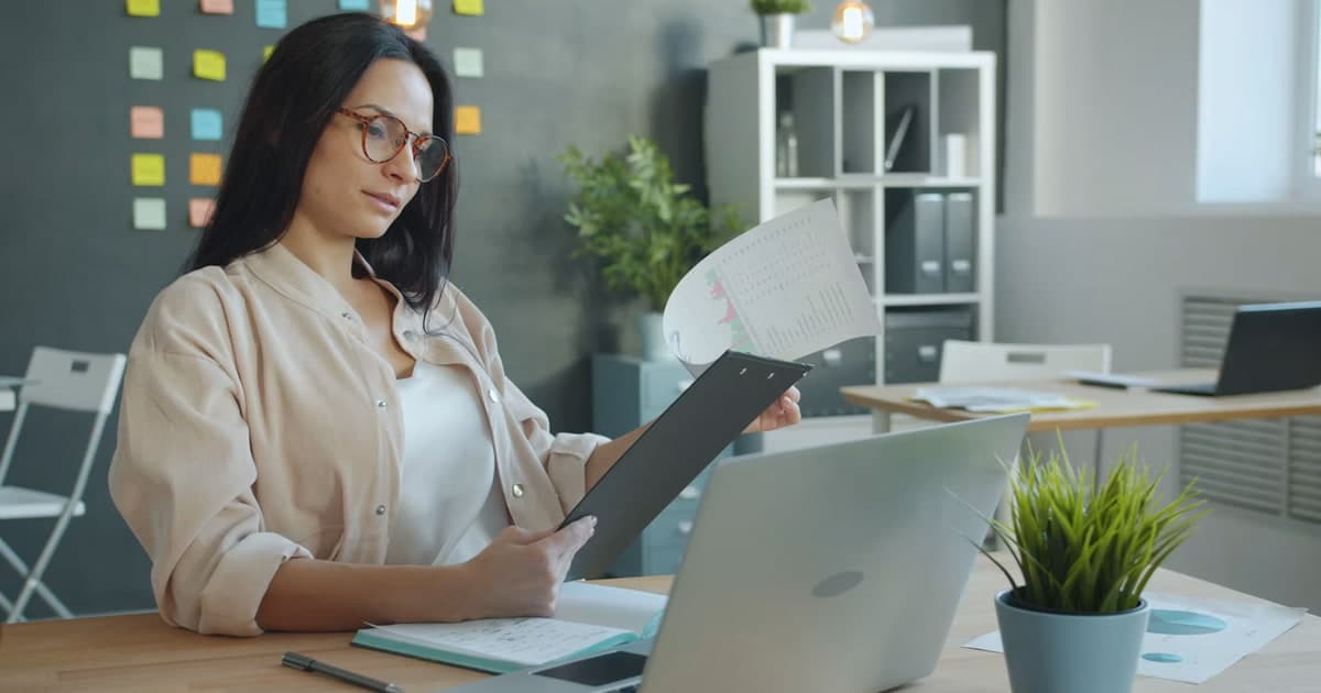 A woman reviews a printed report beside a laptop at an office desk.