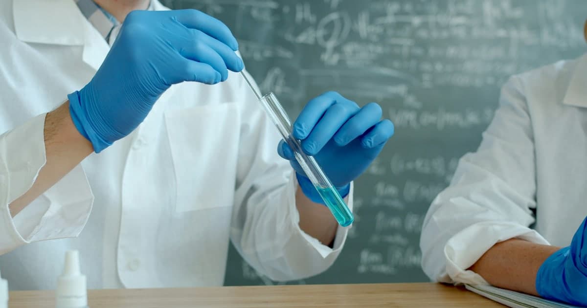 Gloved hands use a pipette to transfer blue liquid into a test tube in a laboratory.