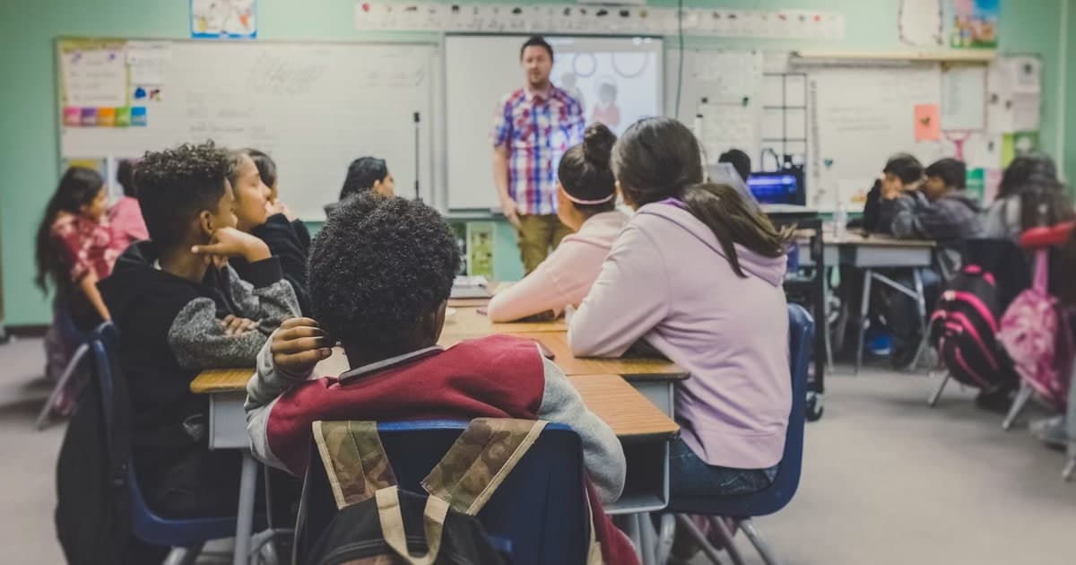 Students sit facing a teacher in a classroom.