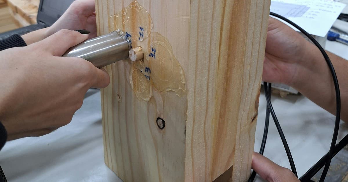 Hands test a wood sample with ultrasonic equipment in a laboratory.