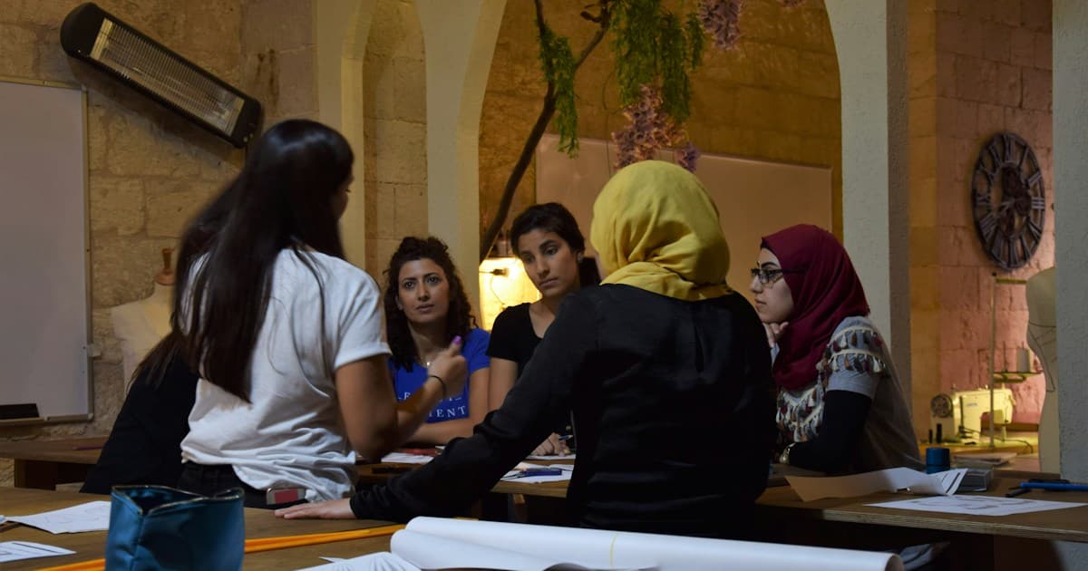 A facilitator speaks with four women seated around a table in a group discussion.