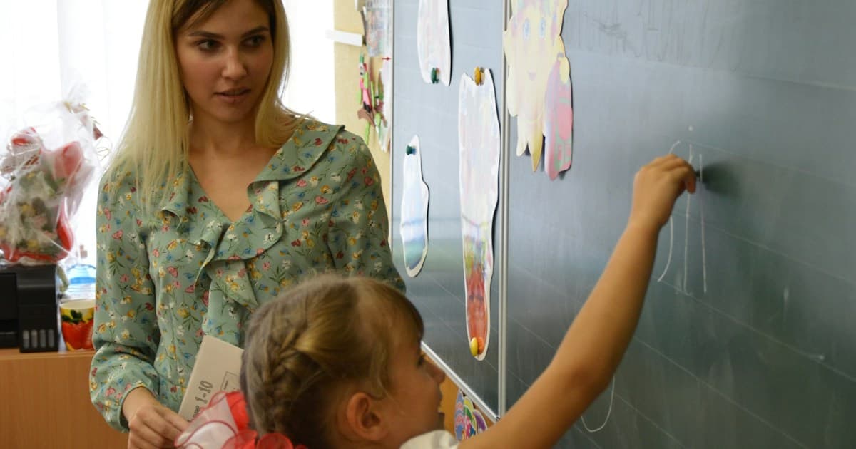 A teacher watches a student write on a classroom board.