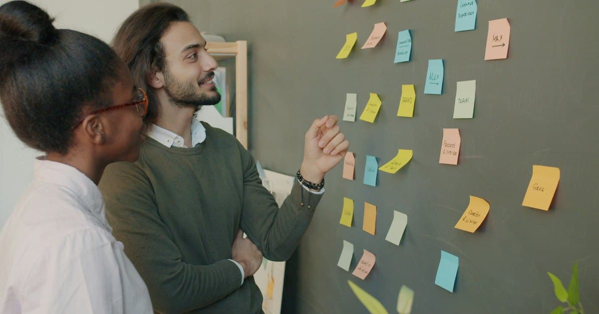 Two colleagues discuss a wall of grouped sticky notes.