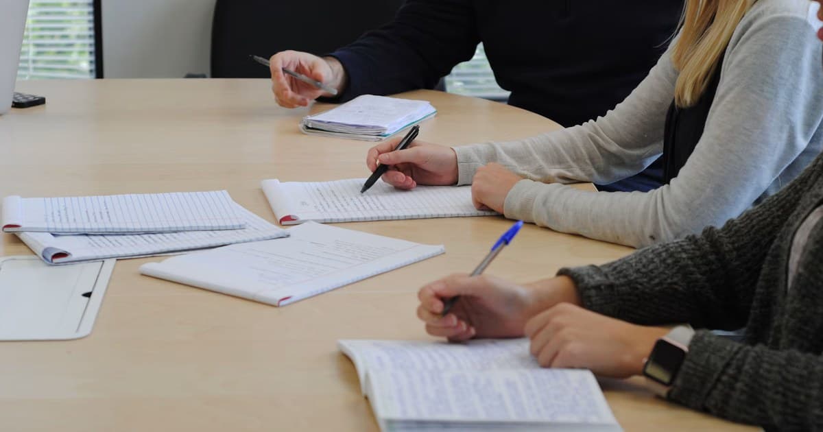 People take handwritten notes in notebooks around a meeting table.