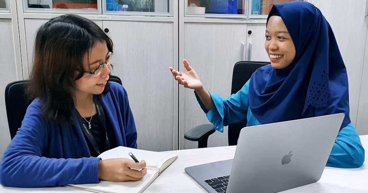 A researcher listens and takes notes as a participant speaks beside a laptop.