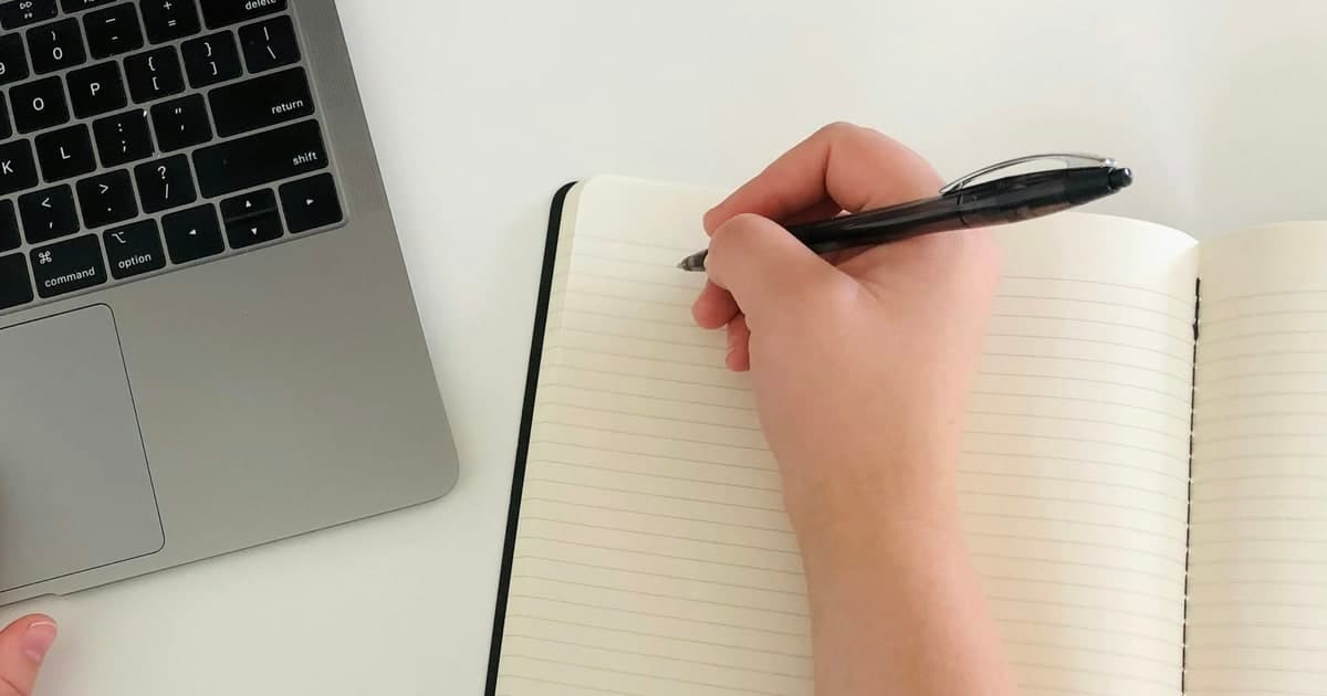 A hand writes in an open notebook beside a laptop keyboard.