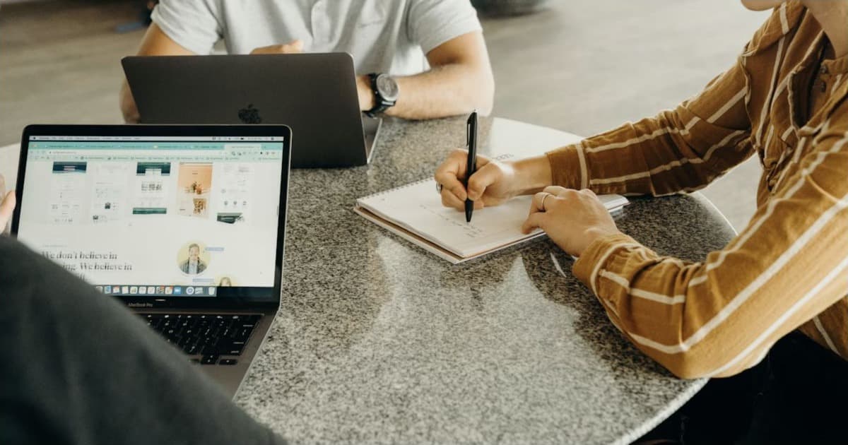Three people meet around a table; one writes in a notebook beside laptops.