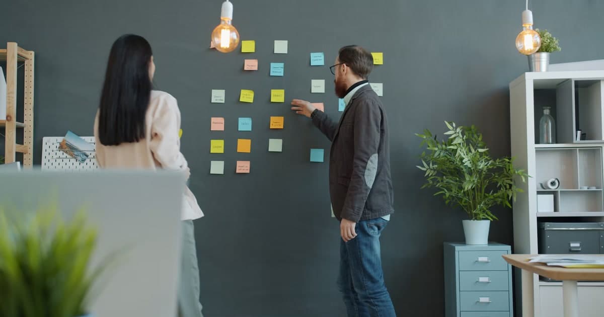 Two coworkers discuss sticky notes arranged across a wall.