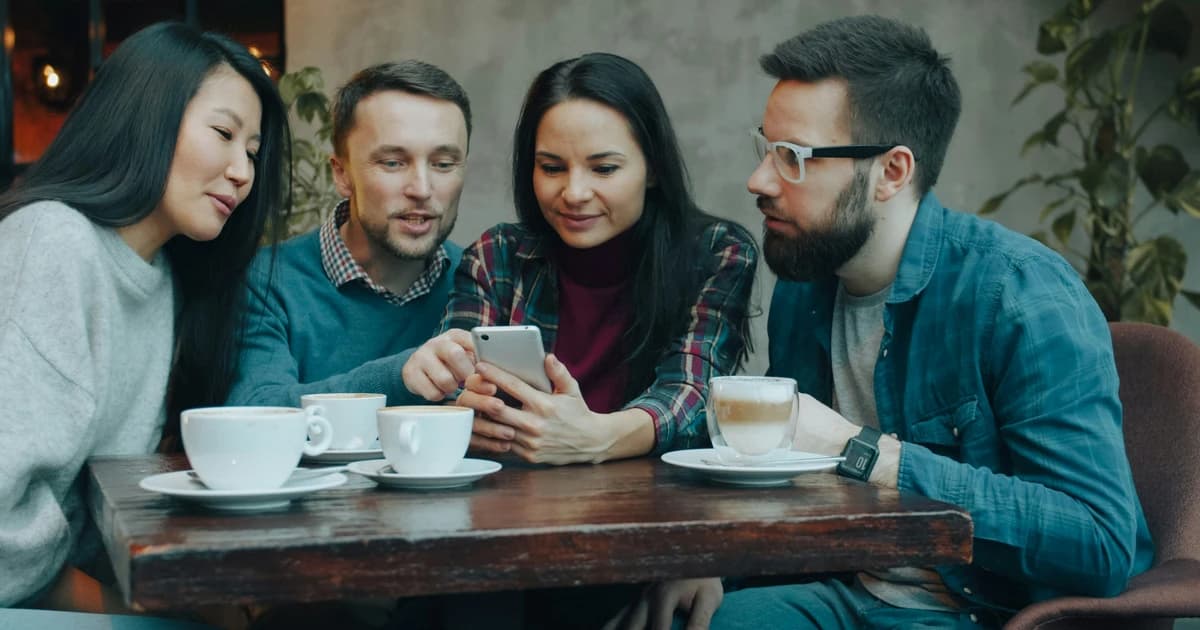 Four people leaning toward a shared smartphone at a cafe table.