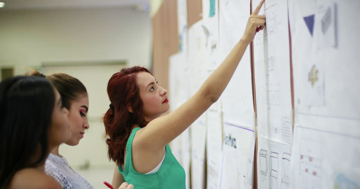 A woman points to wall diagrams while two others look on and take notes.