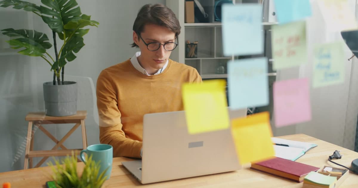A person works at a laptop beside notebooks and wall sticky notes.