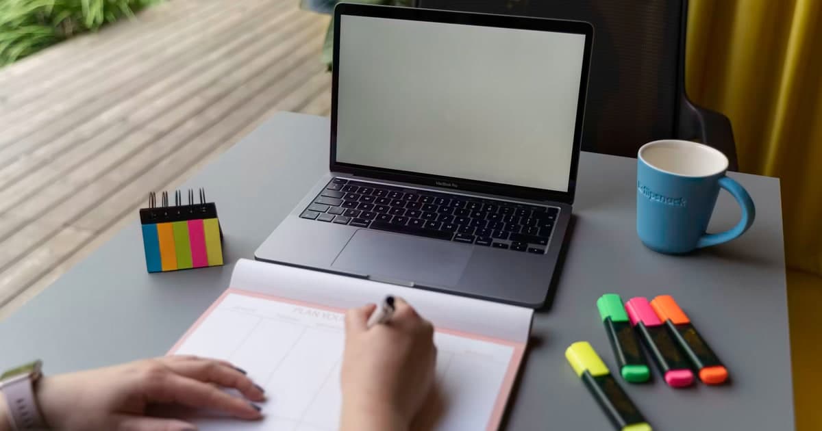 A person writes a plan beside a blank laptop, sticky notes, and markers.