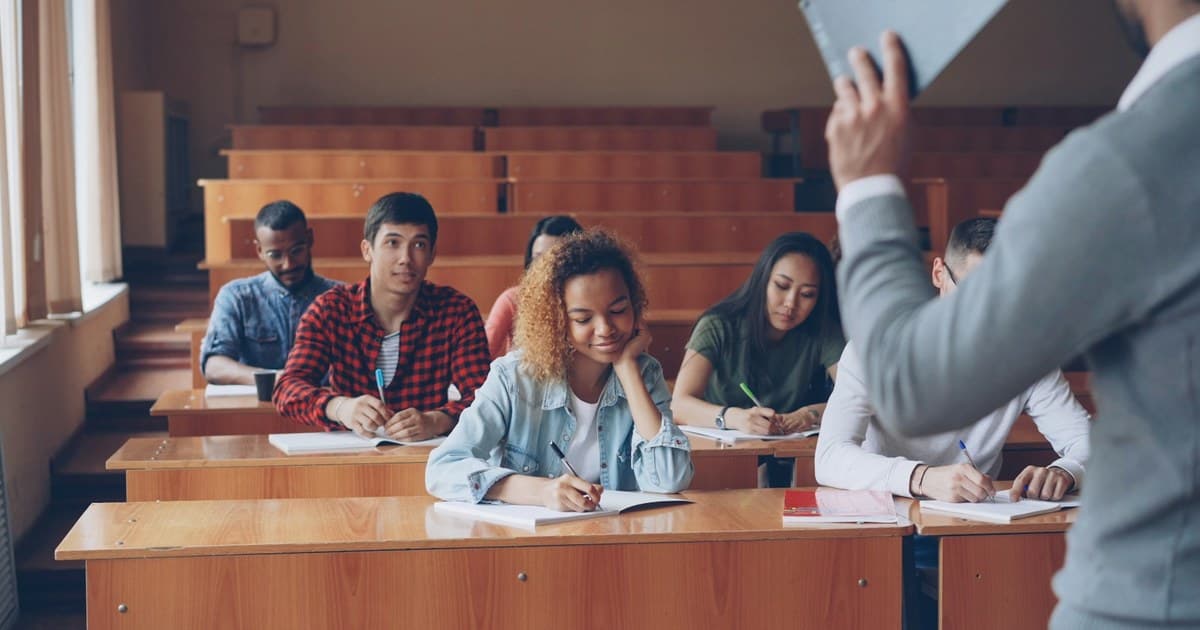 Students write notes as an instructor speaks at a classroom lecture.