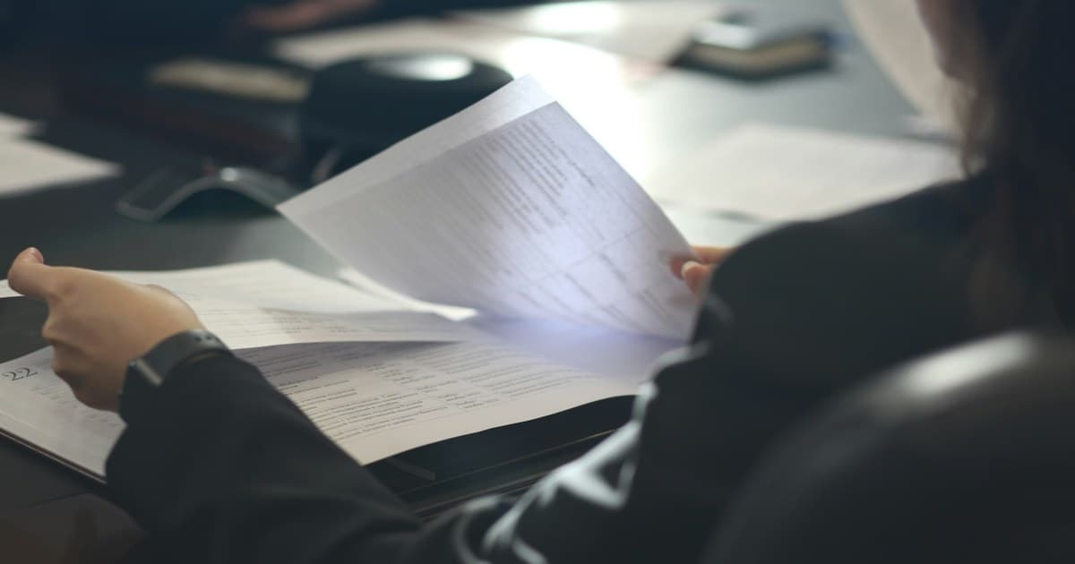 A person reads a printed page above several other papers at a table.