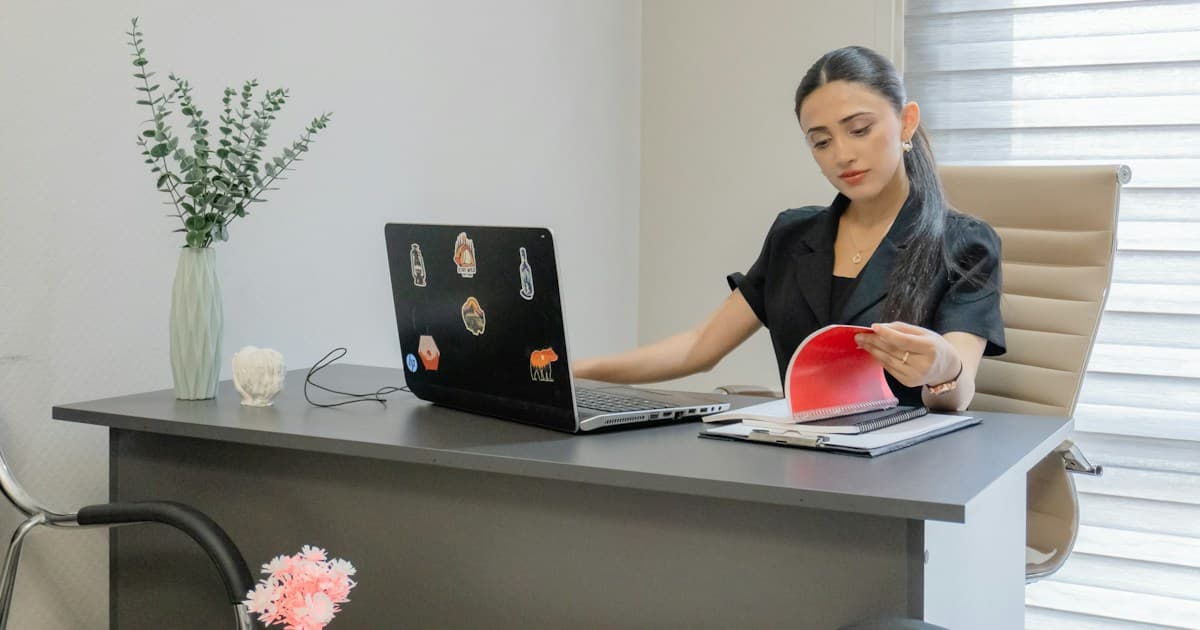 A person reviews a notebook beside an open laptop at a desk.