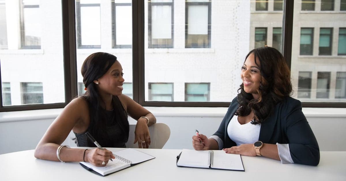Two women talk across a table while holding pens and notebooks.