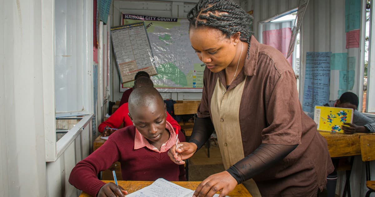 A teacher works beside one student at a classroom desk.