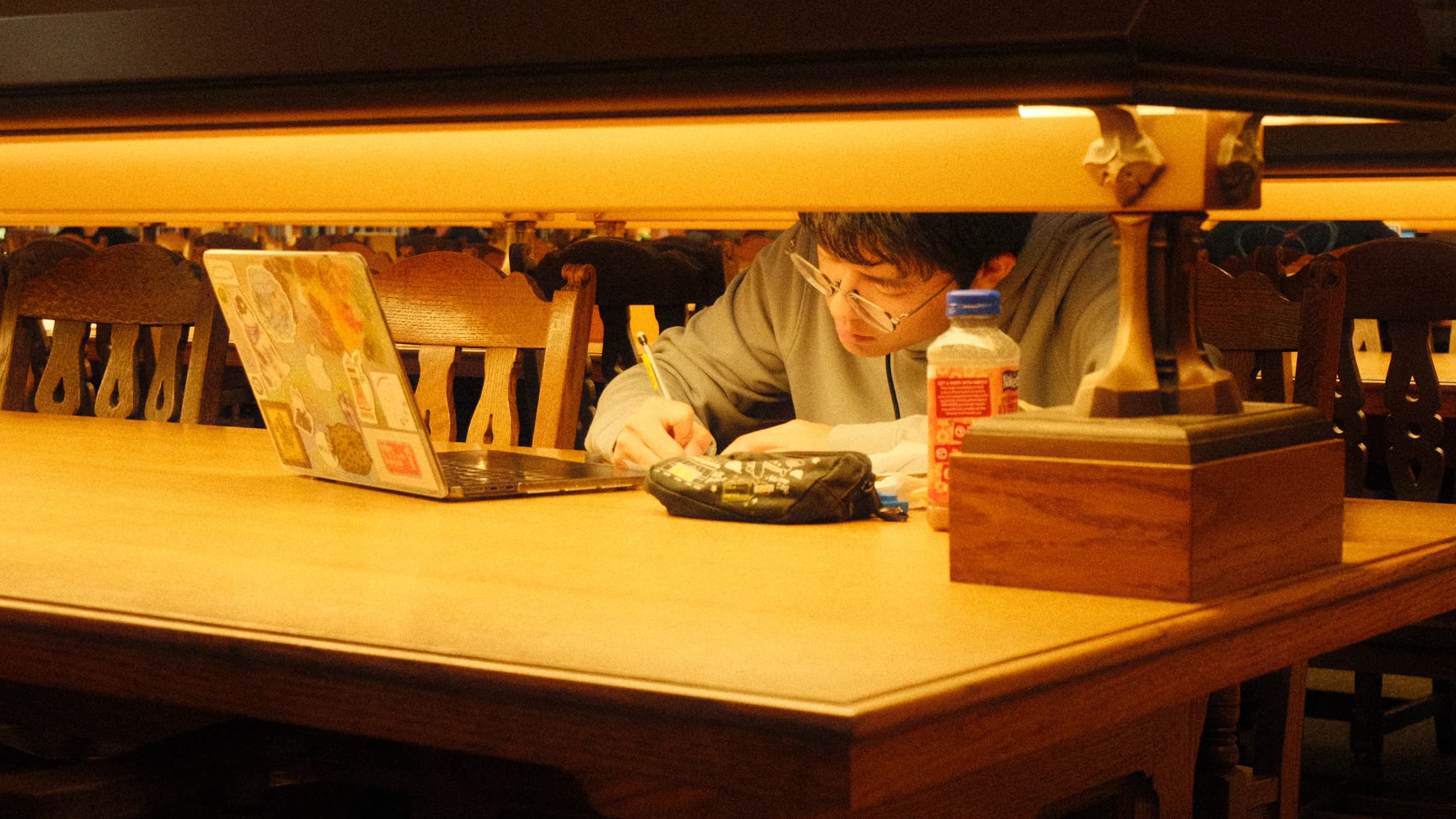 A student reads and writes beside a laptop at a library table.
