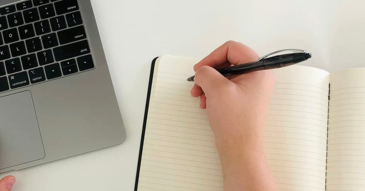 A hand writes in an open notebook beside the edge of a laptop keyboard.