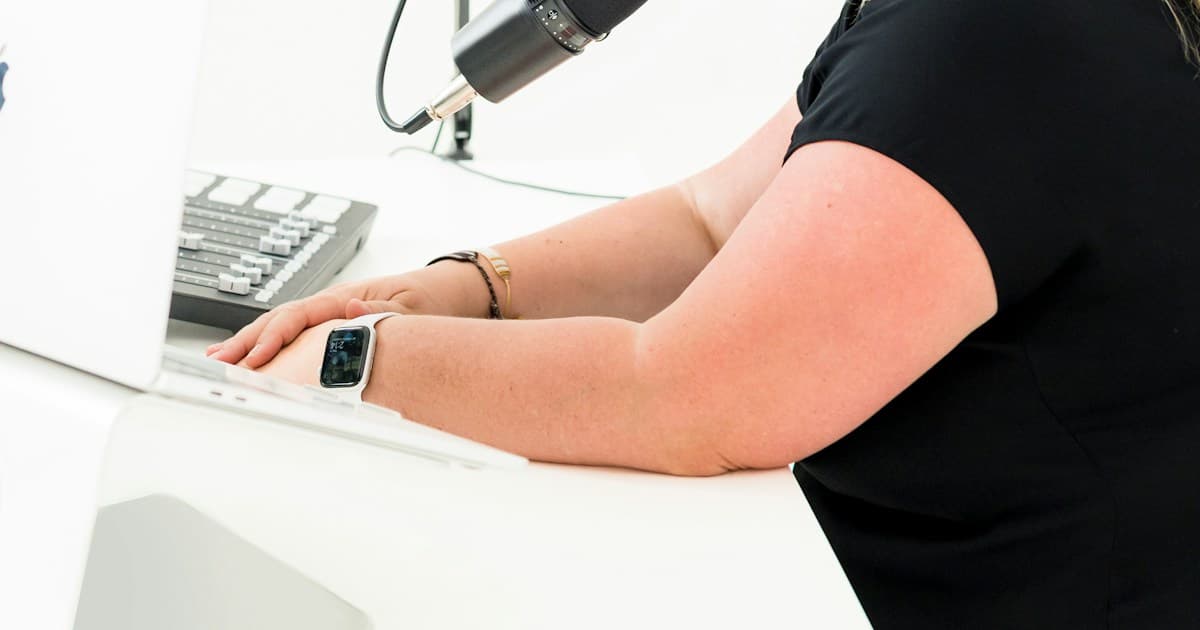 A person works at a laptop beside a podcast microphone while wearing headphones.
