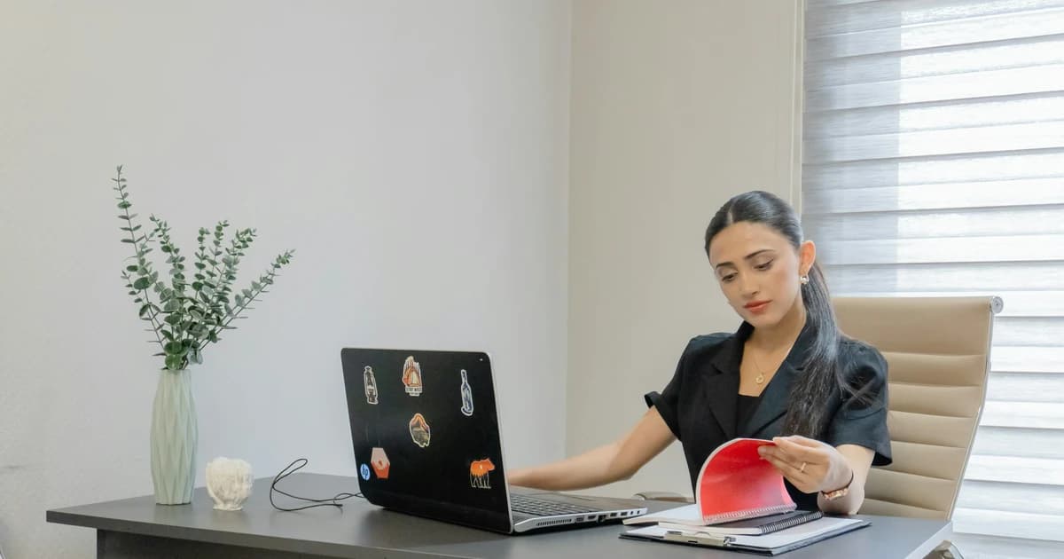 A woman reads a printed page beside an open laptop.
