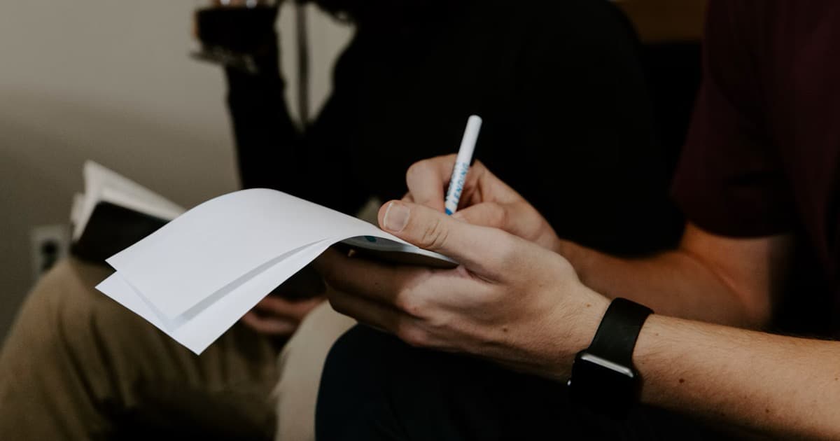 A person writes notes on paper beside another seated person.