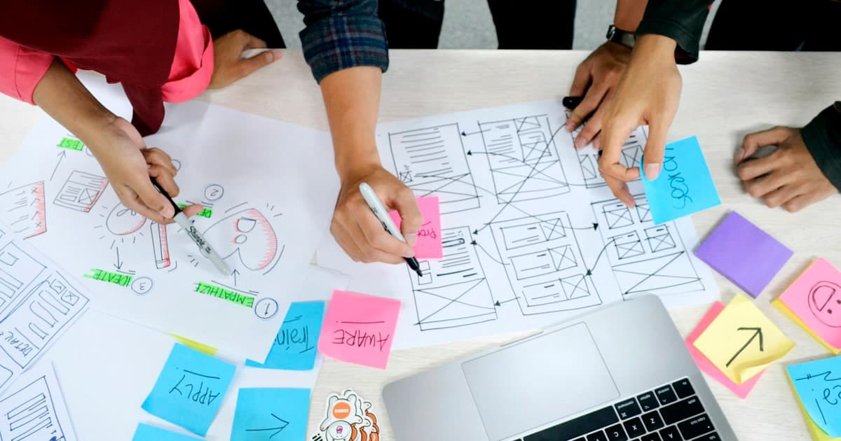 A team sketches web-page wireframes on paper beside a laptop and sticky notes.